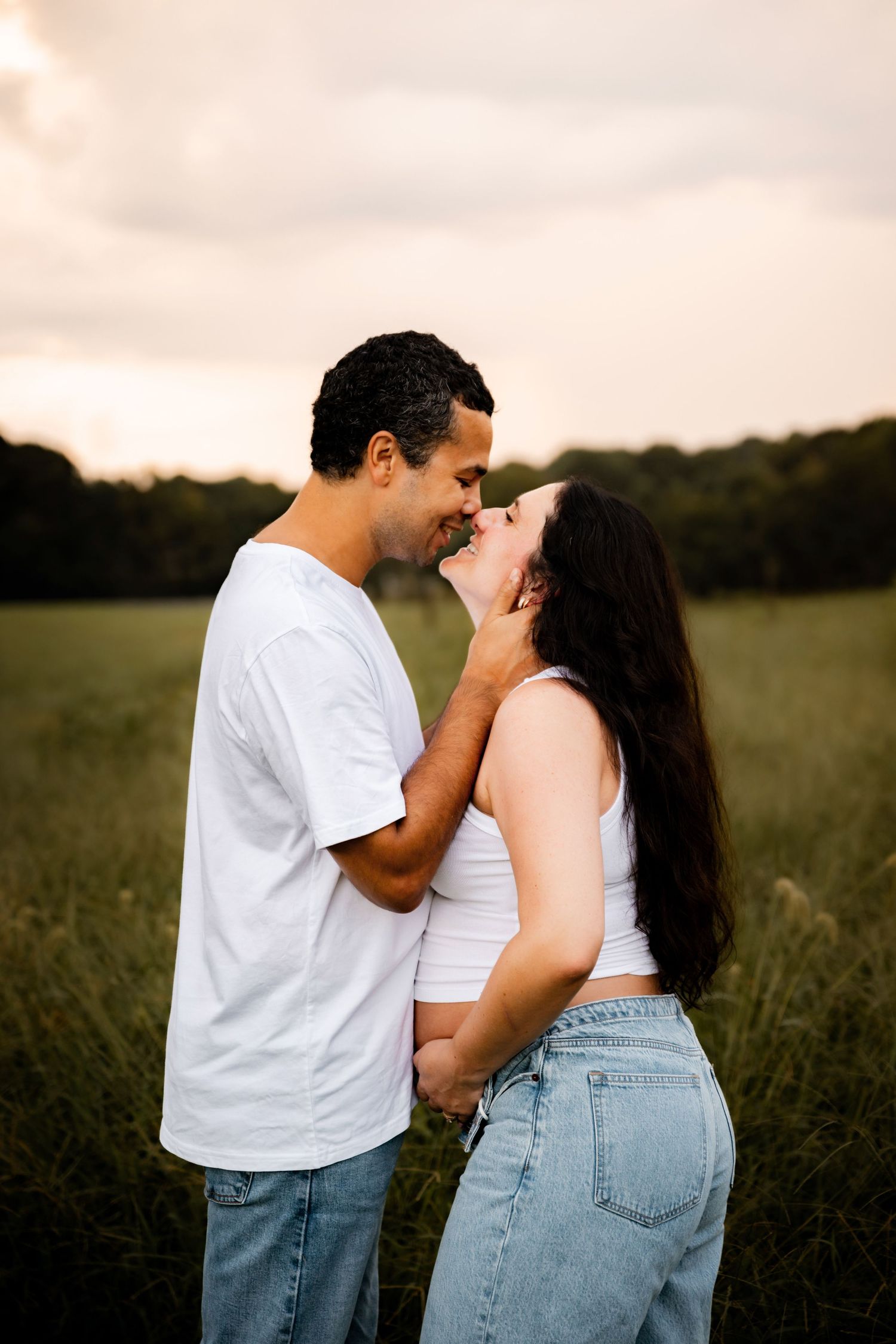 Couple in matching white tops and jeans sharing a romantic moment in a green field at golden hour sunset.