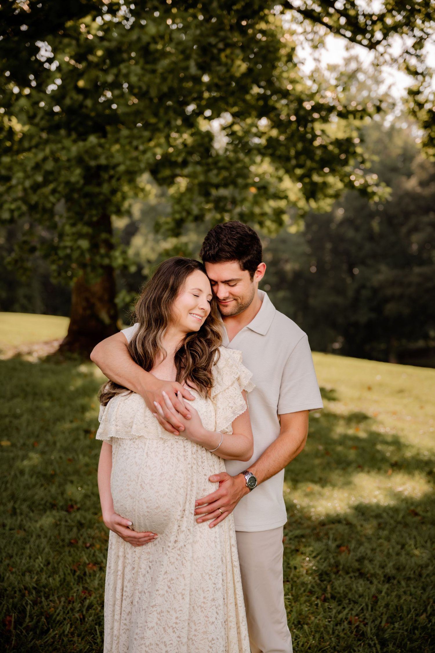 Couple shares tender maternity photo outdoors, man embracing pregnant woman in white dress under lush green trees in golden light.