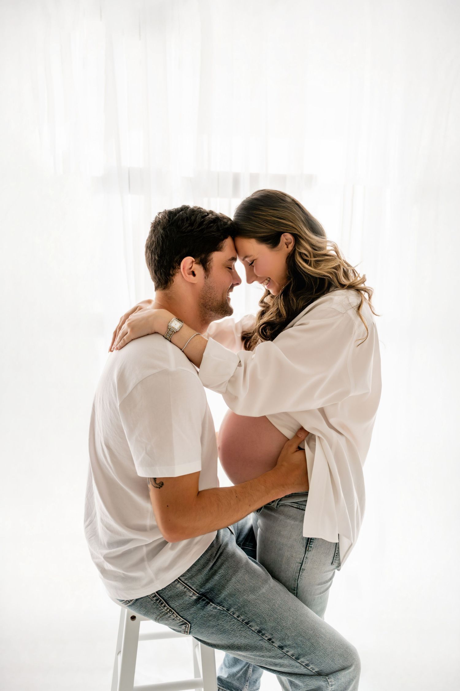 A couple shares an intimate moment, with a pregnant woman in an open white shirt embracing a man seated on a stool.