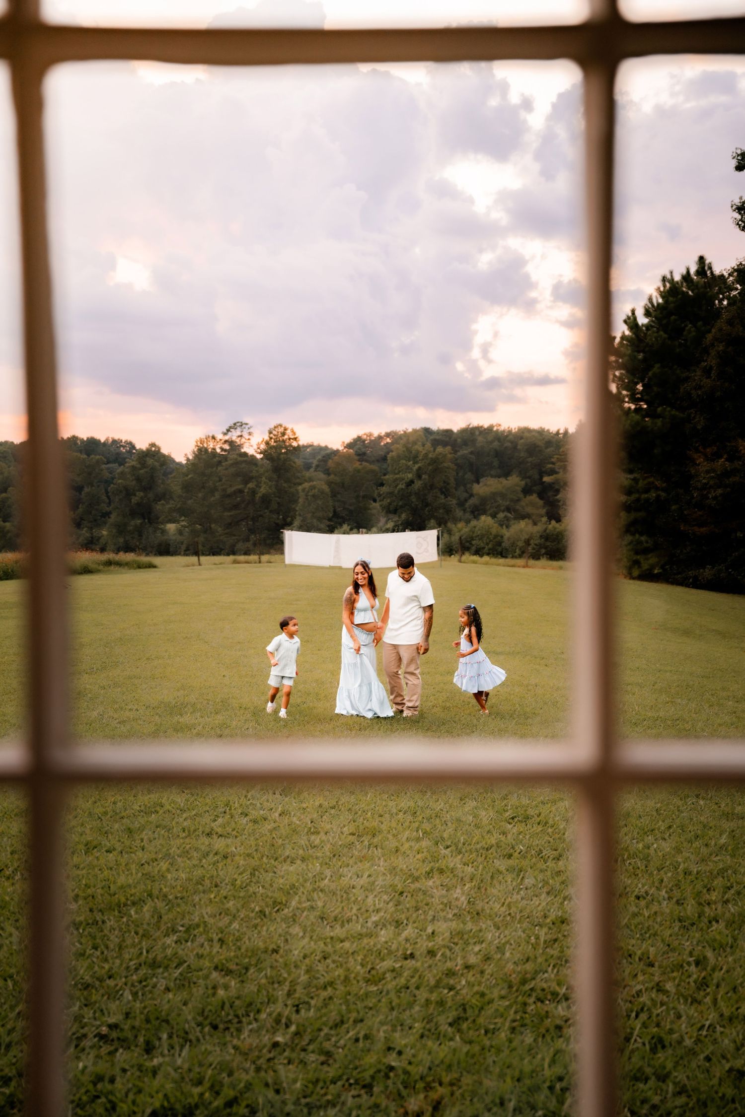 Family of four in white outfits standing in a green field at sunset, framed through a window.