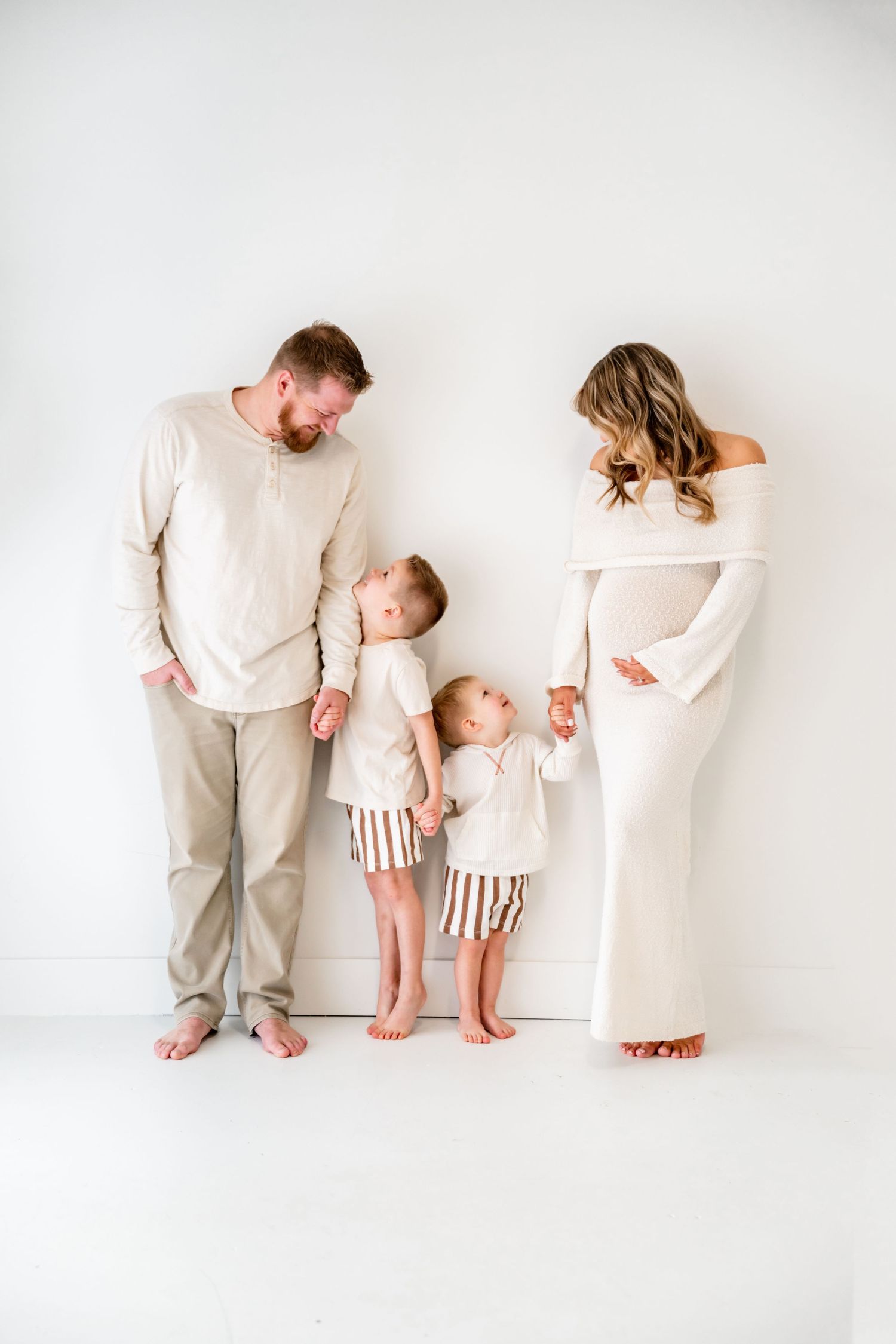 Family wearing coordinated neutral outfits stands together against white wall in minimalist studio setting.
