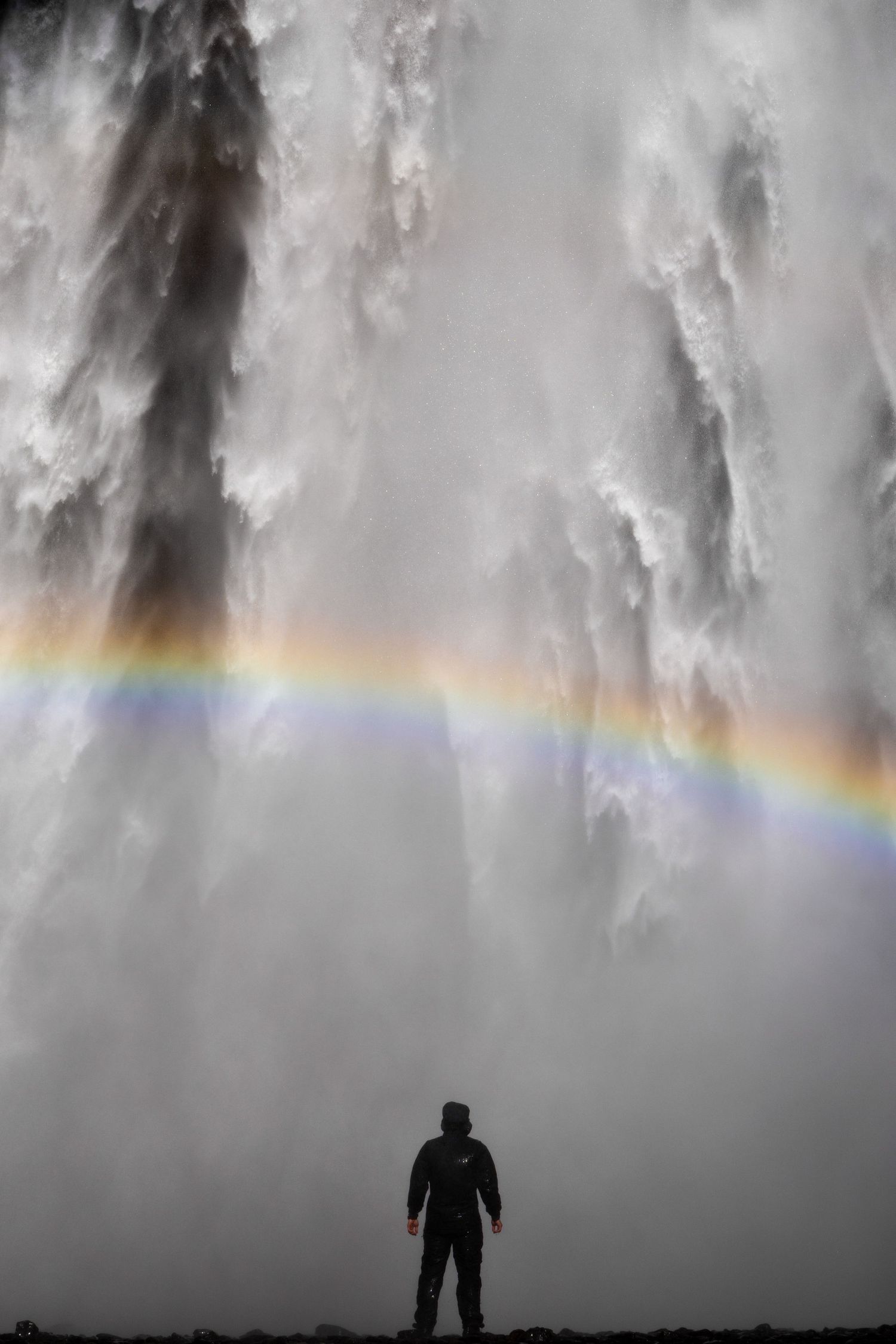 Silhouetted figure stands beneath dramatic gray clouds with vibrant rainbow stretching across misty sky.