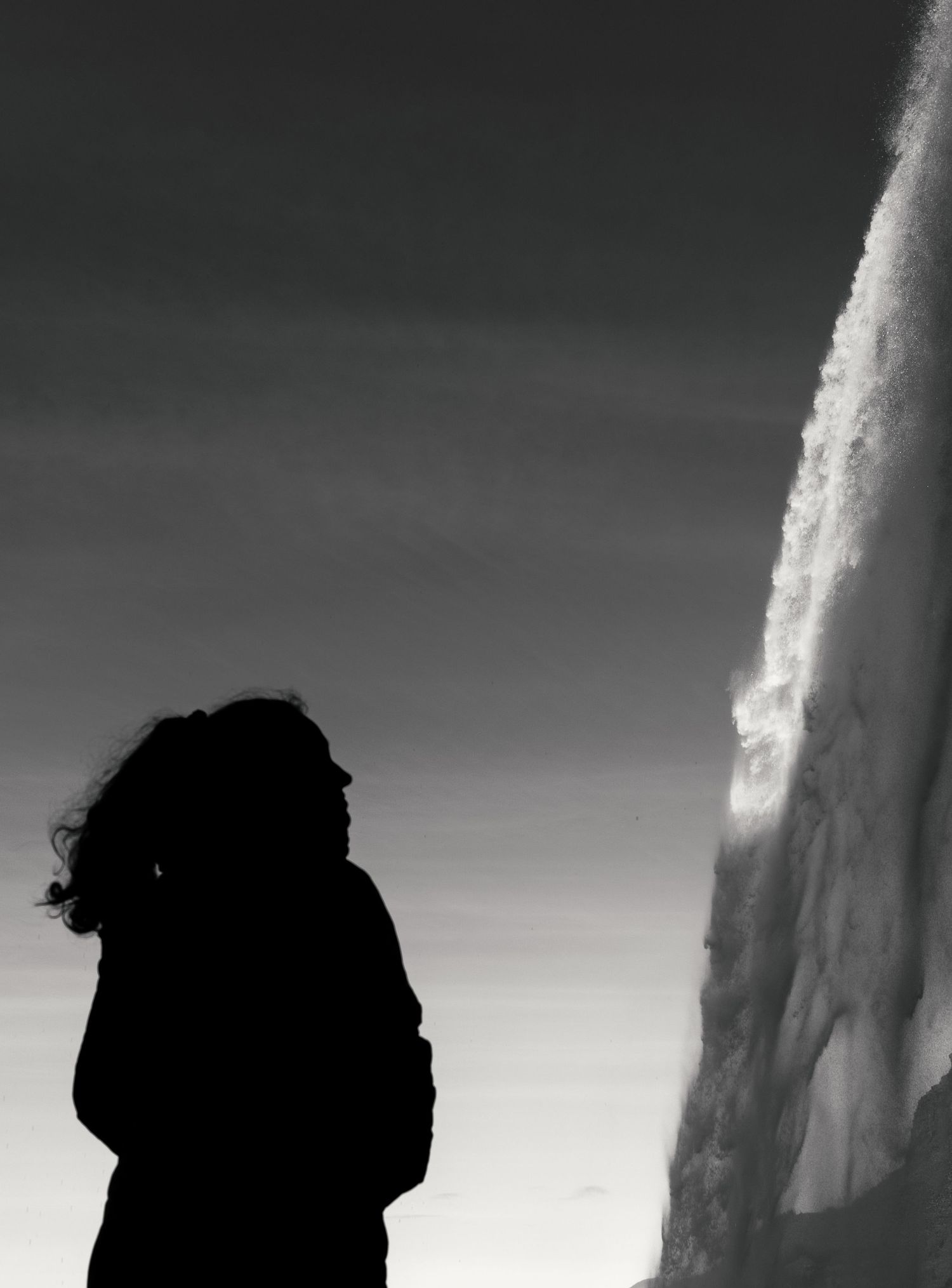A silhouetted figure stands against a dramatic waterfall backdrop in black and white photography.