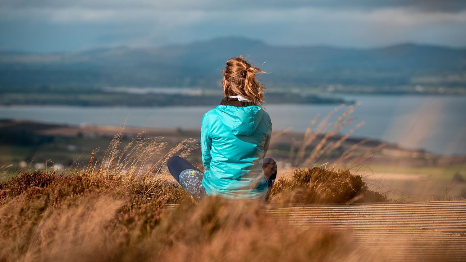 Person in turquoise jacket sits peacefully on hilltop overlooking mountain lake landscape in soft evening light.