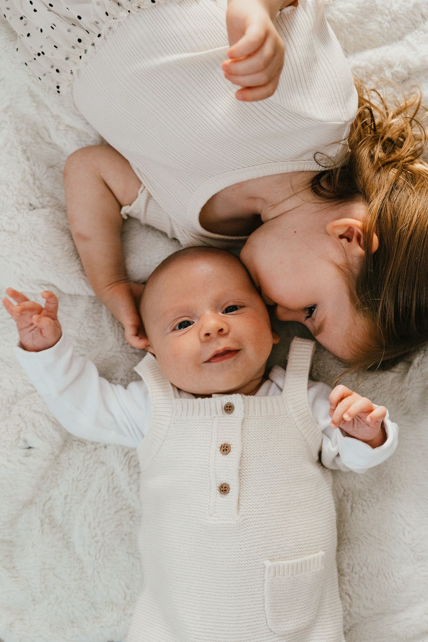 Baby wearing white sweater lying on textured blanket while being kissed on a soft white bed.