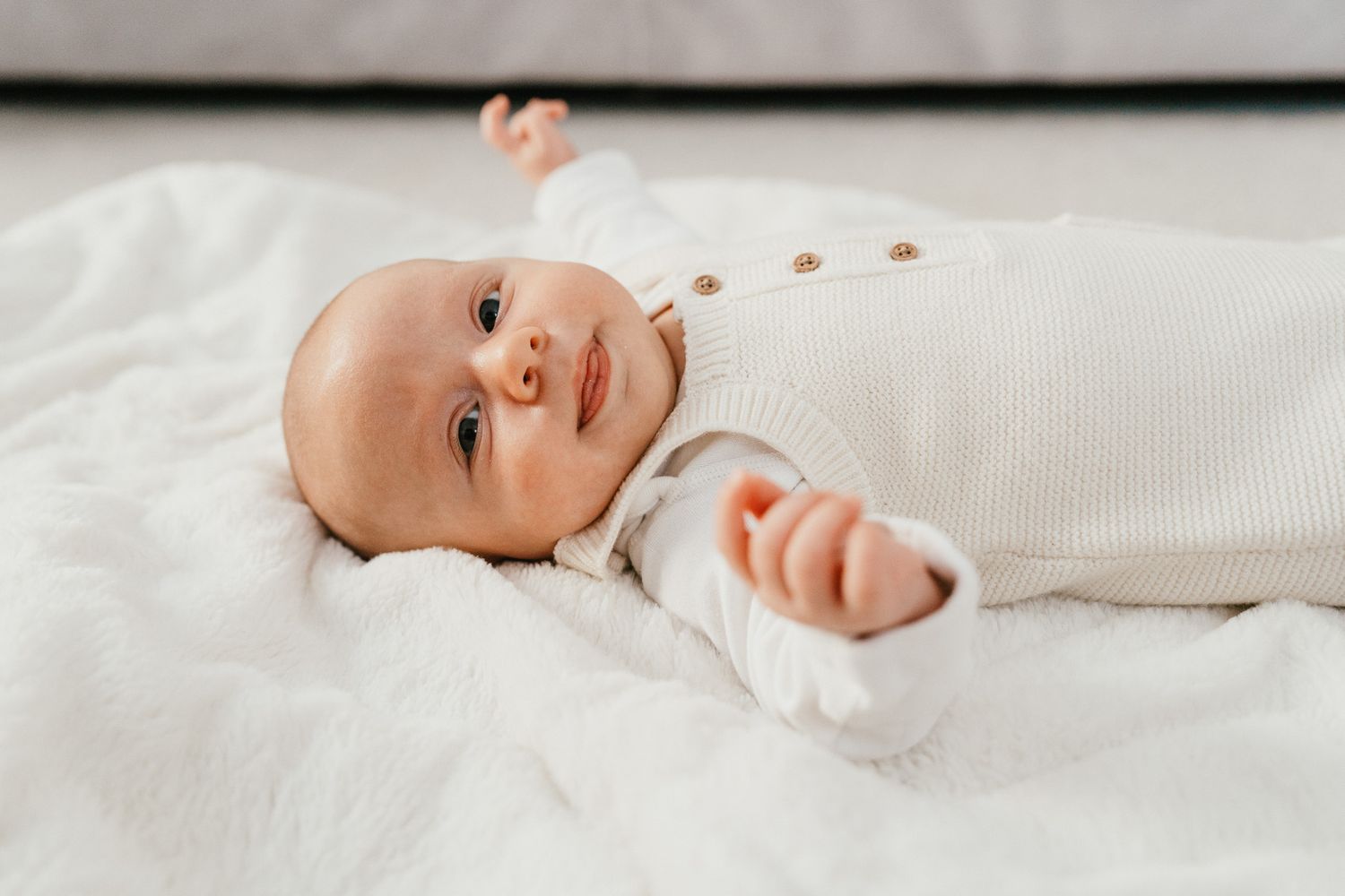 Baby wearing white onesie lying on white bedding looking up at camera.
