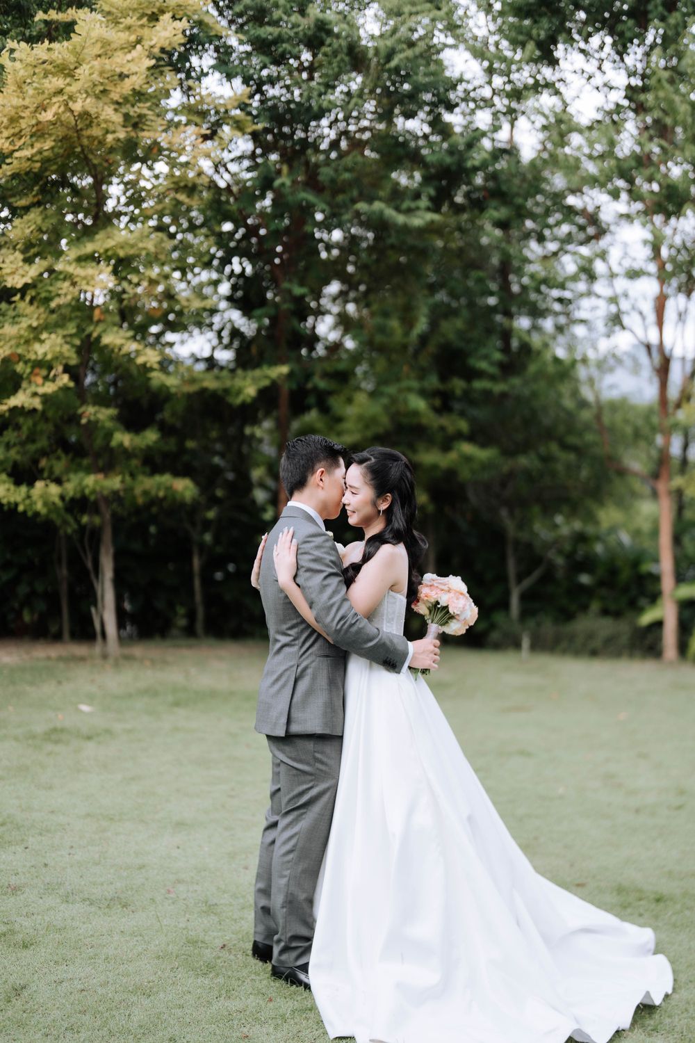 A romantic wedding couple embraces on a lush lawn surrounded by trees during their outdoor ceremony.