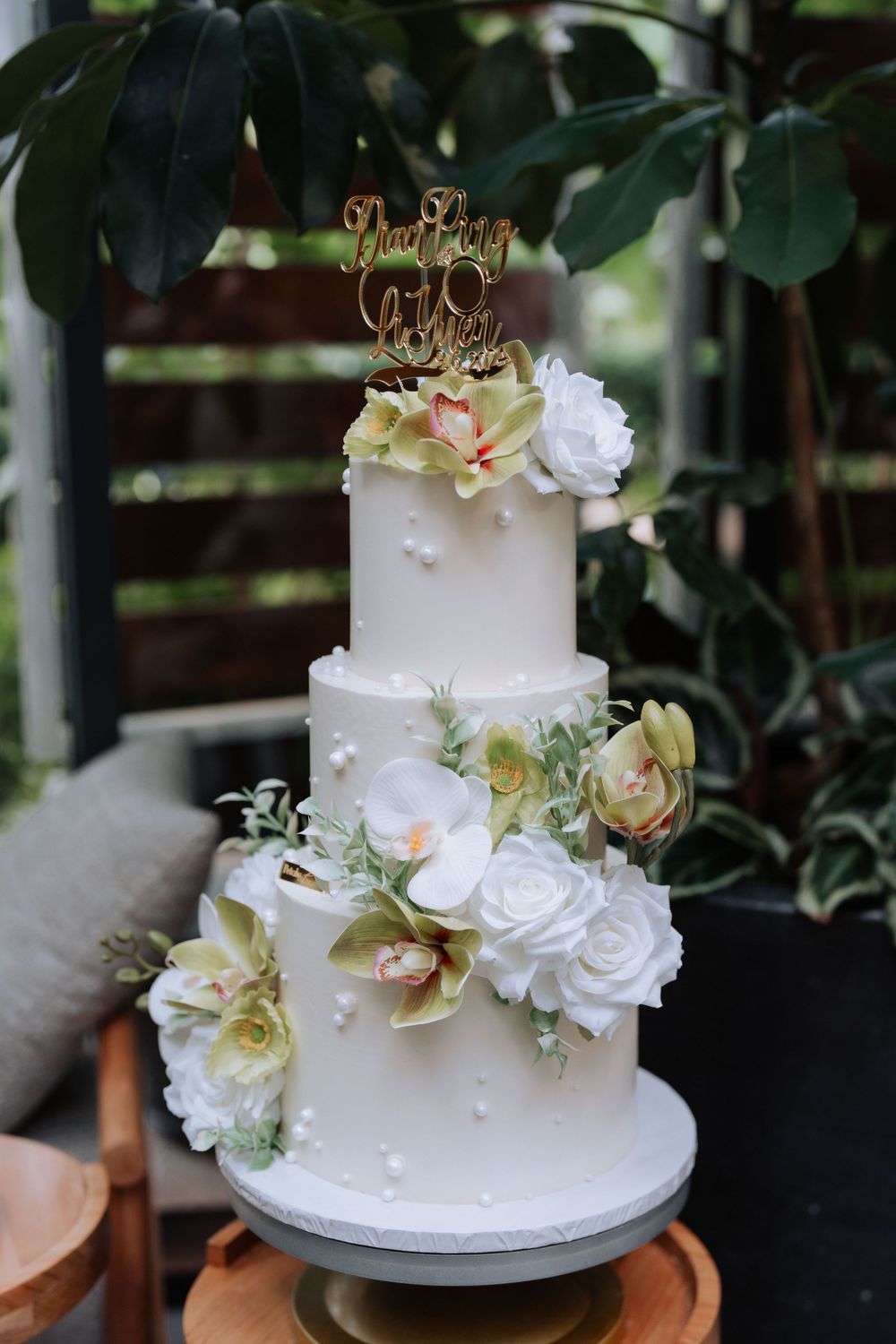 Elegant three-tier white wedding cake decorated with white roses and orchids against a garden backdrop.