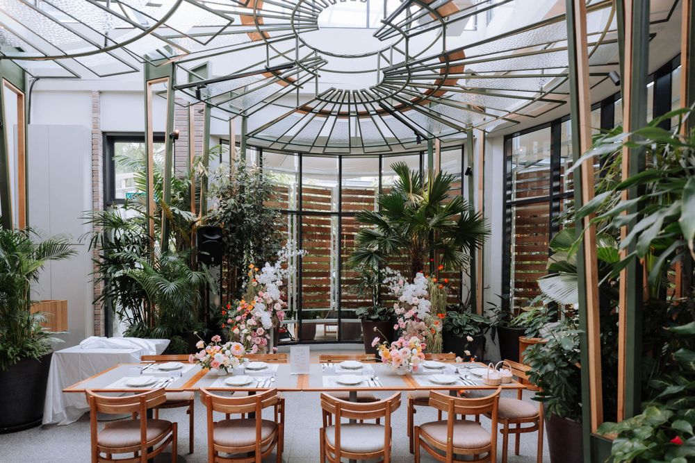 Greenhouse wedding reception setup with wooden chairs and floral decorations under a glass ceiling.