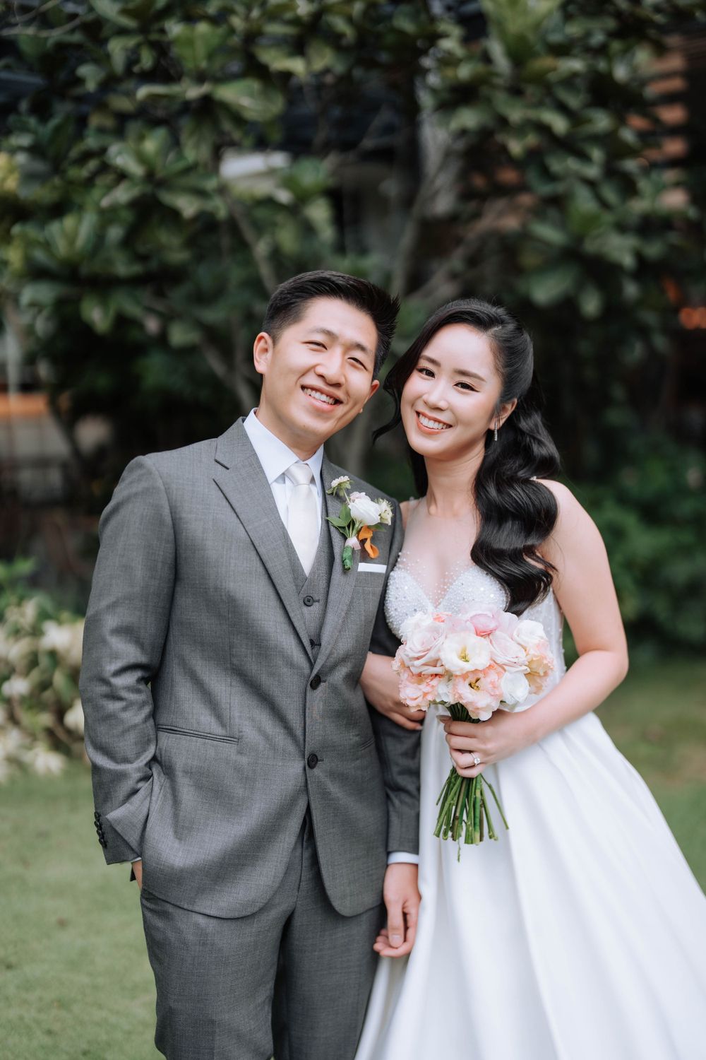 A happy wedding couple stands together on grass with trees in the background.