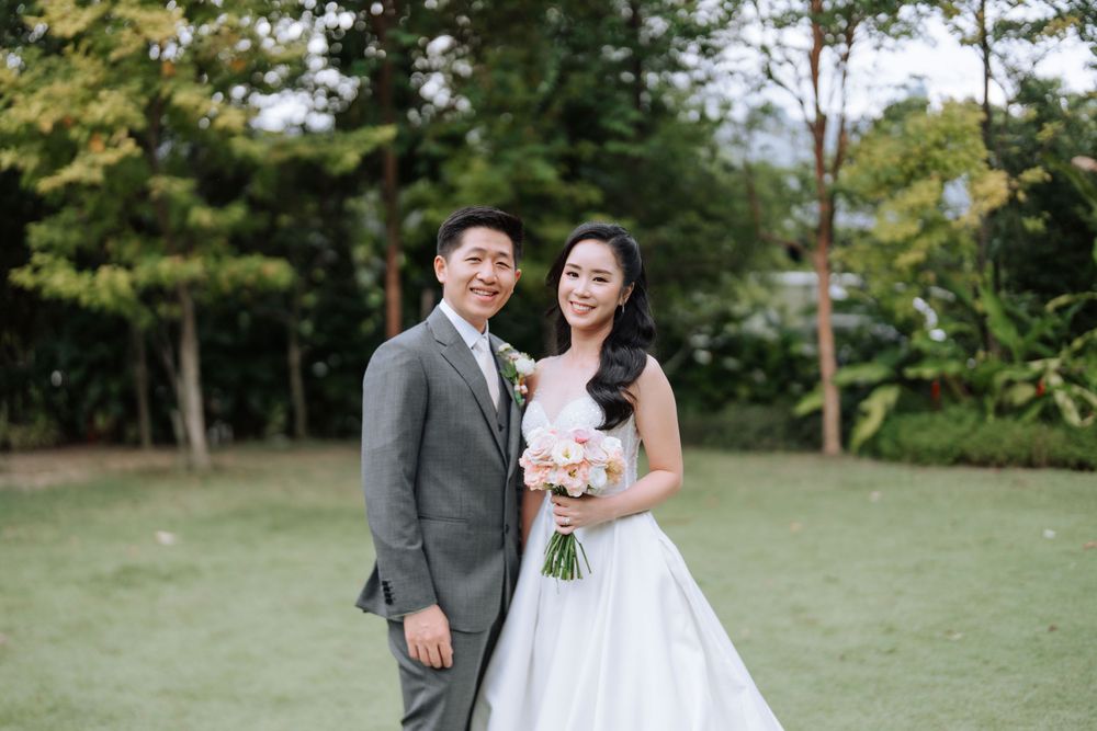 A wedding couple poses together on a lawn with trees in the background.