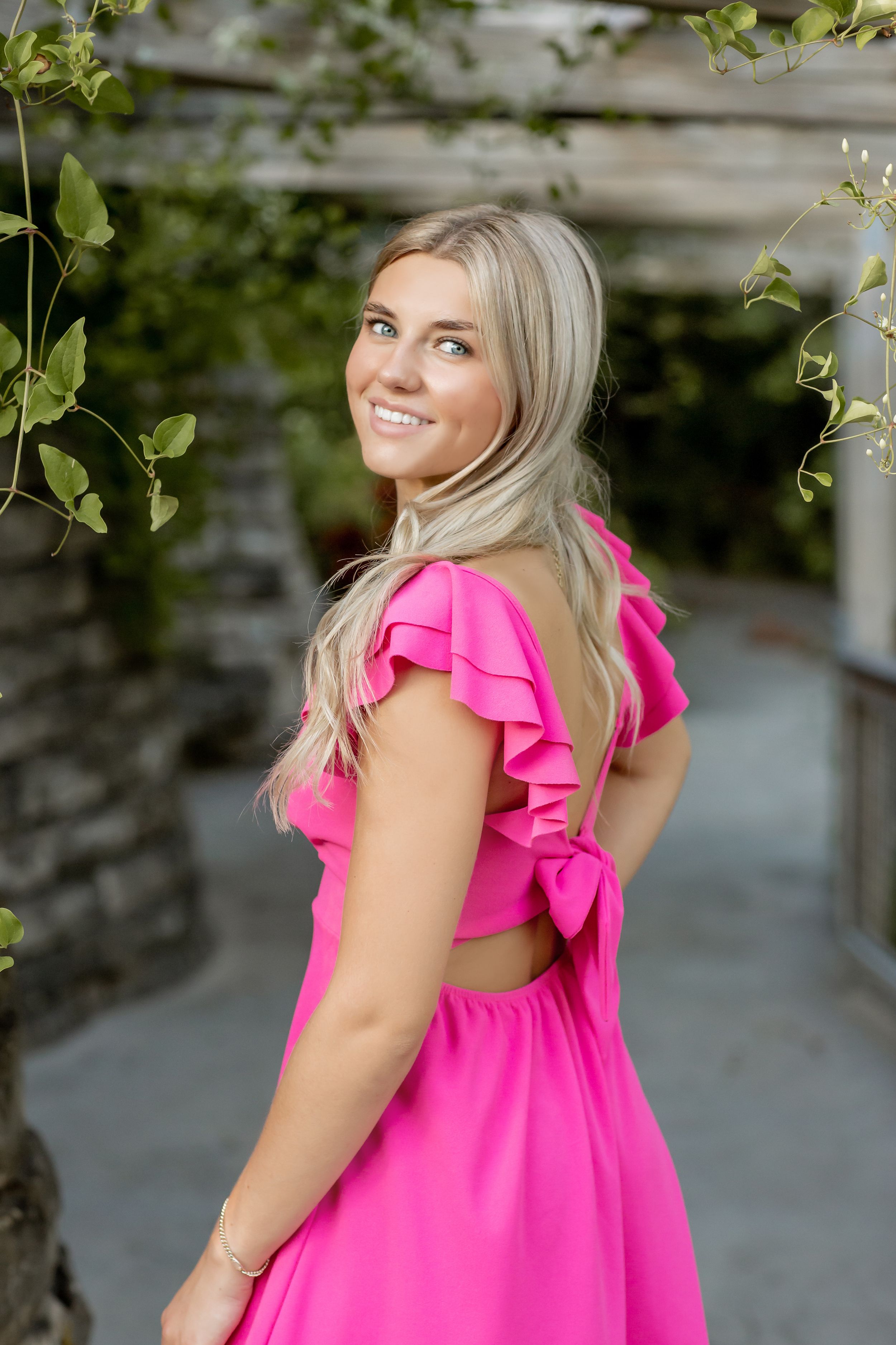 Smiling blonde woman in pink ruffled backless dress posing in a lush garden setting.