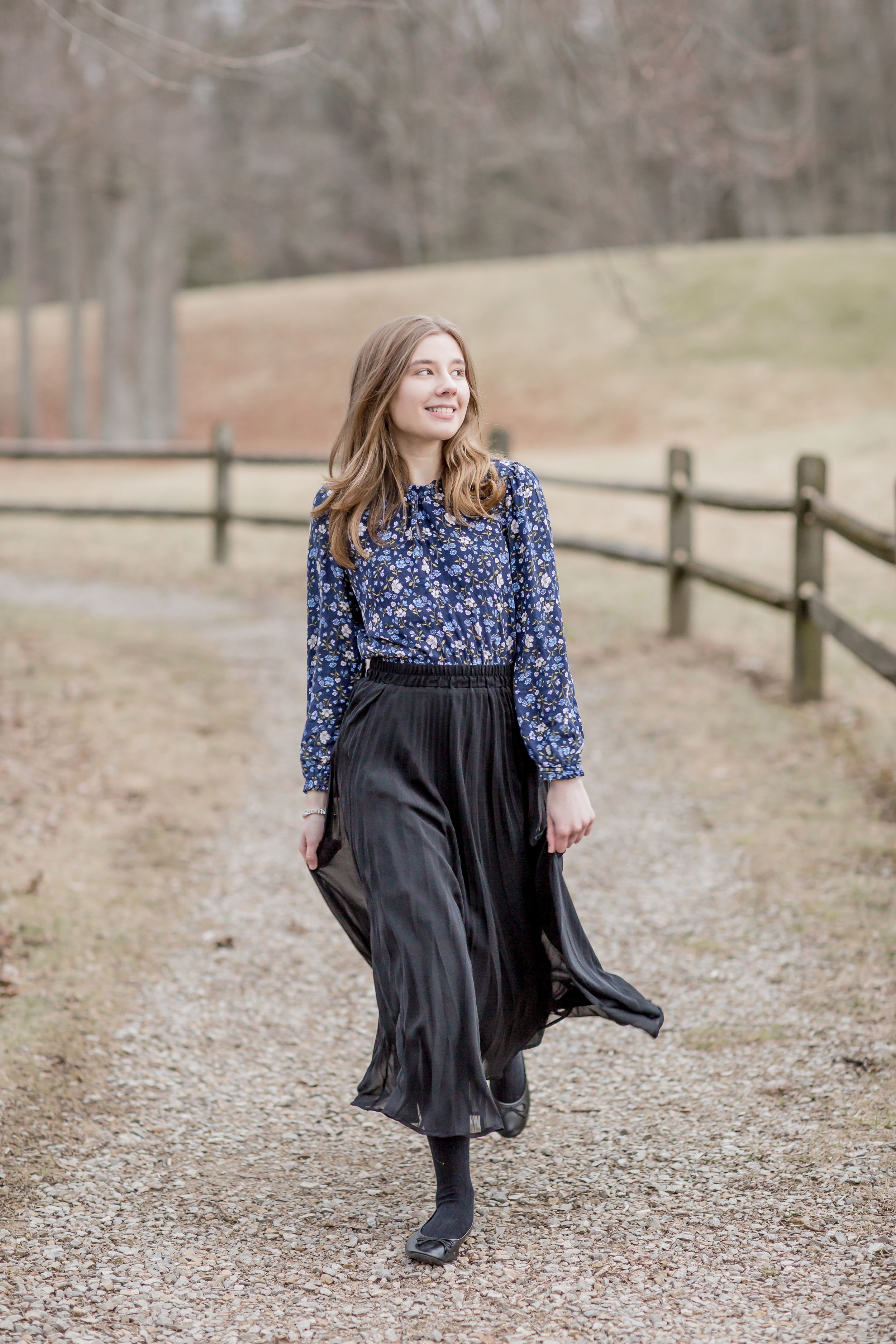 Young woman in blue floral blouse and black skirt walking on a gravel path by a fence.