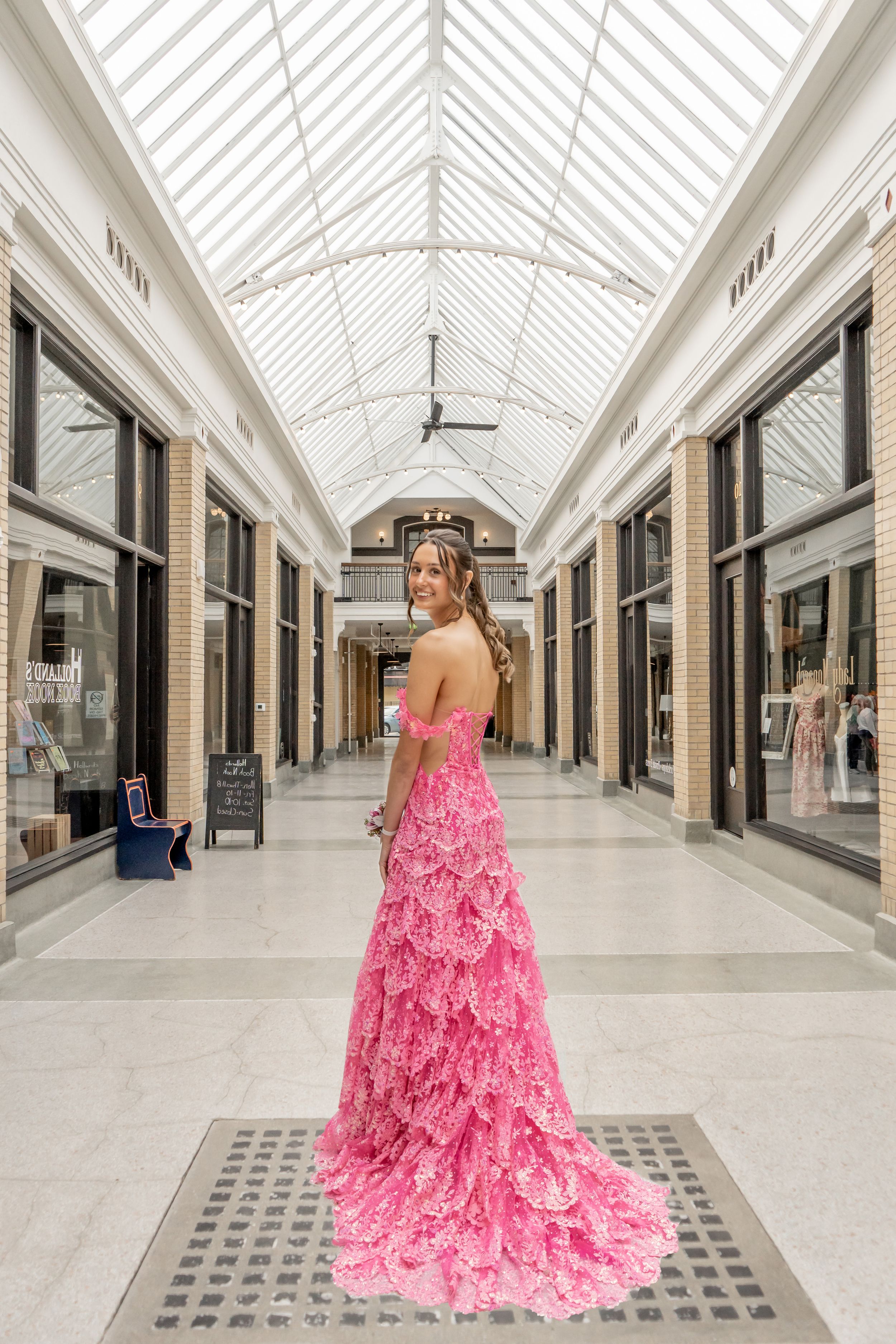 Young woman in elegant pink ruffled prom gown posing in a bright indoor shopping arcade.