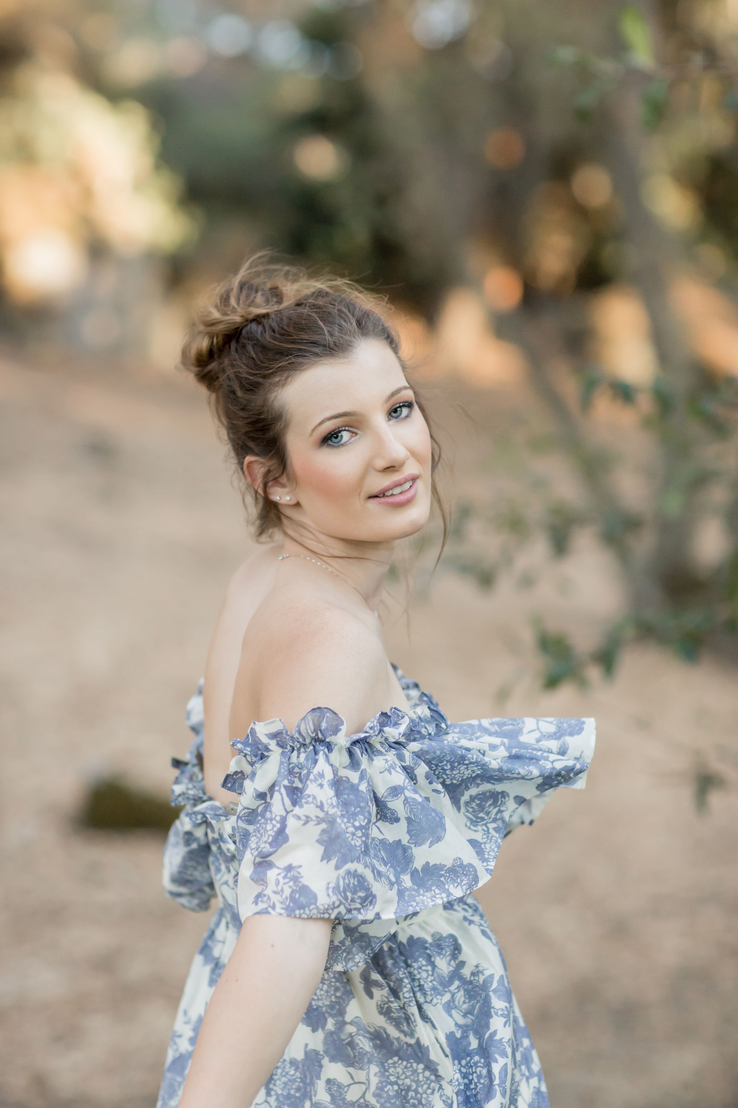 Young woman in blue floral off-shoulder dress glancing back in a sun-dappled outdoor setting.