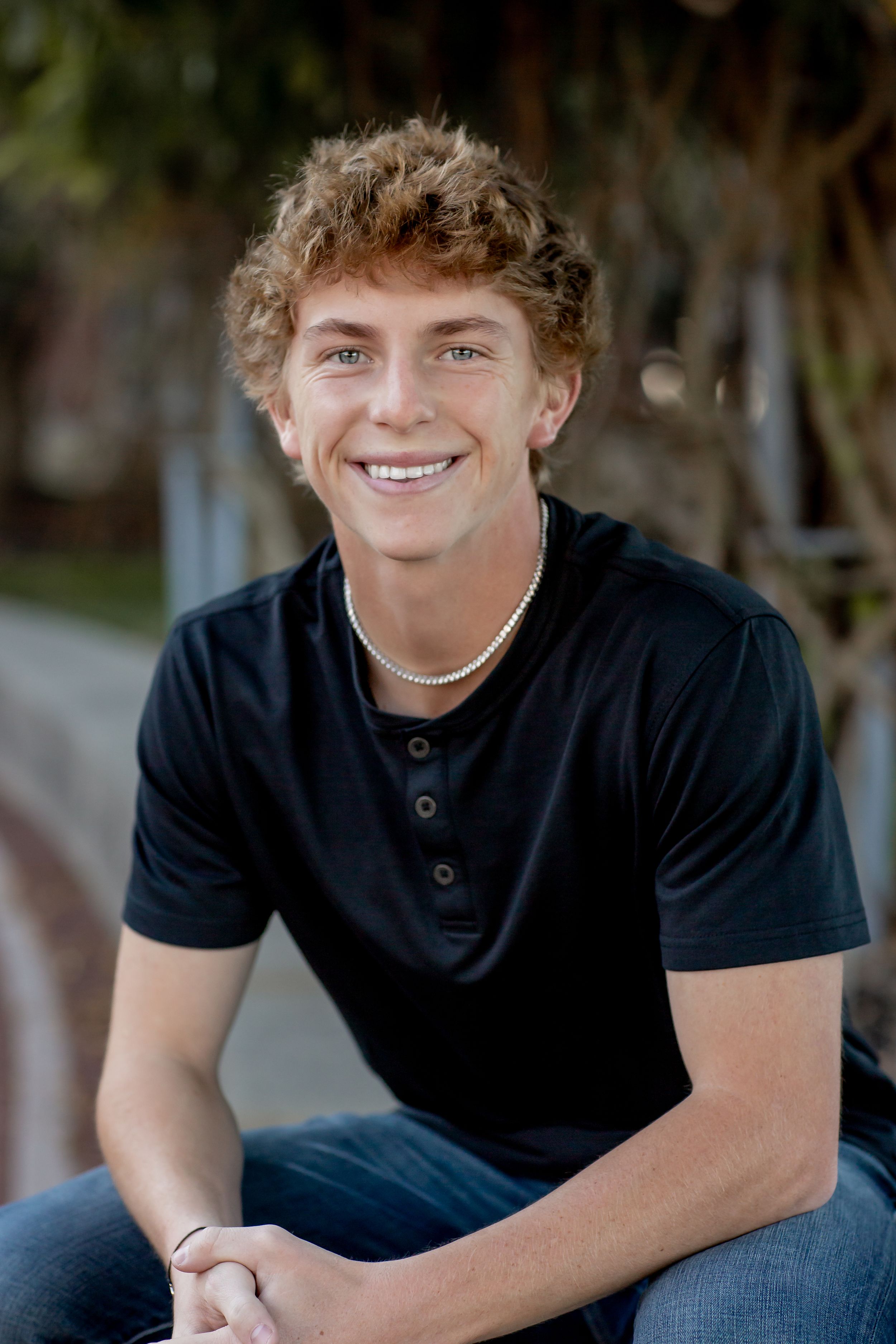 Smiling teen boy with curly hair wearing black shirt and pearl necklace seated outdoors.