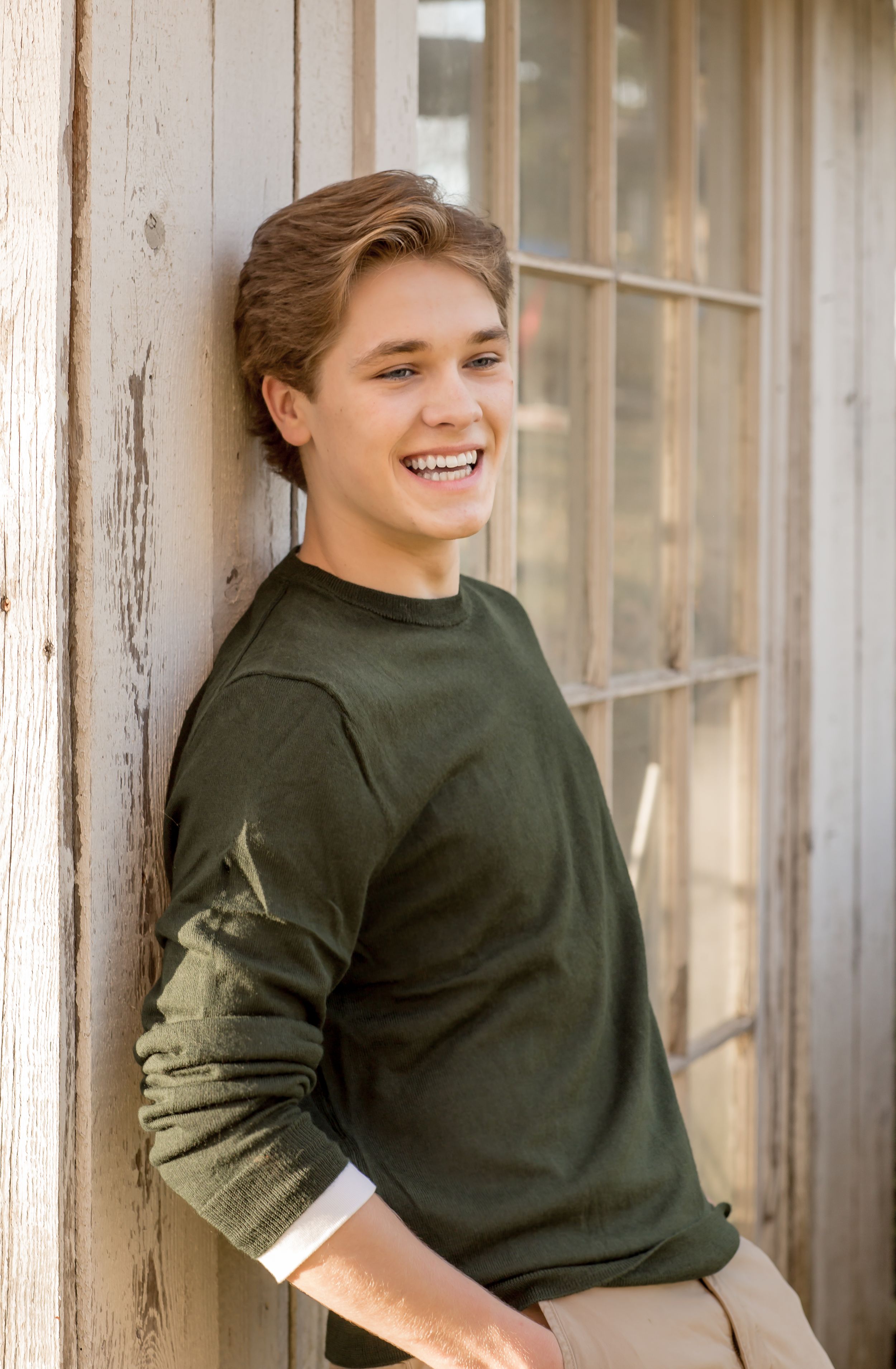 Laughing teen boy in olive green sweater leaning against rustic wooden wall outdoors.
