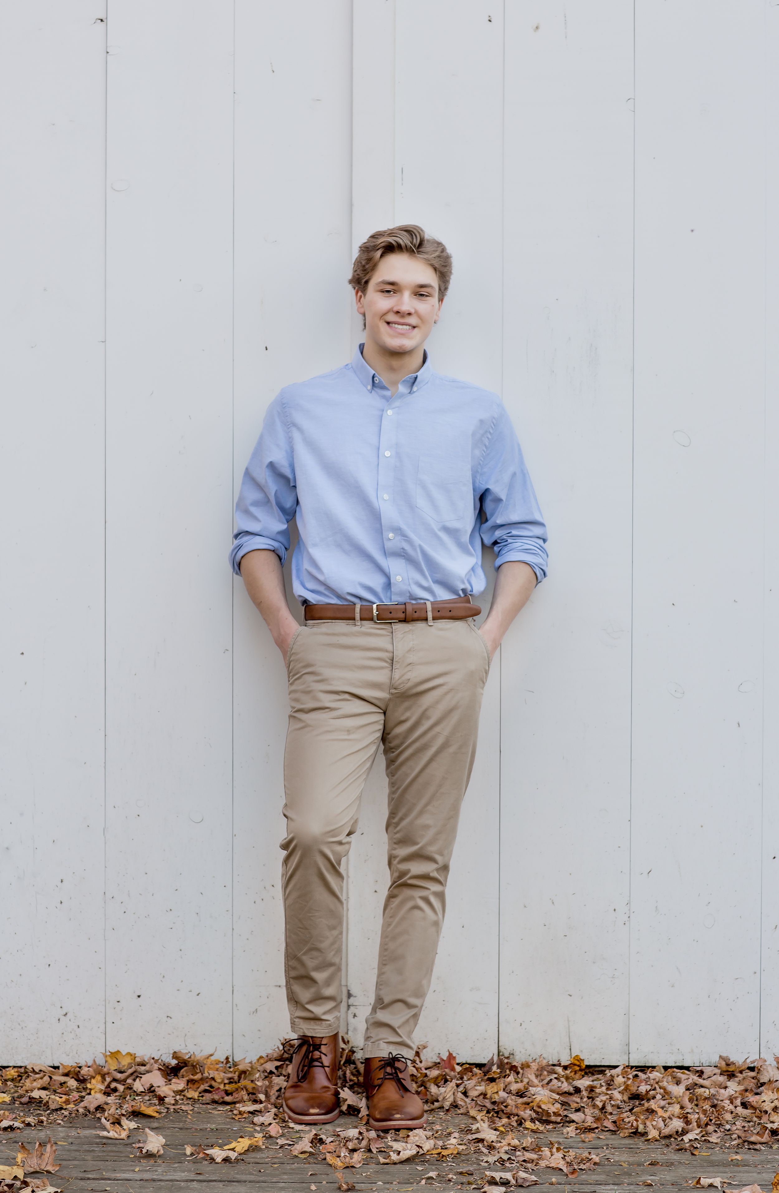 Smiling young man in blue oxford shirt and khakis leaning against white wall with fall leaves.