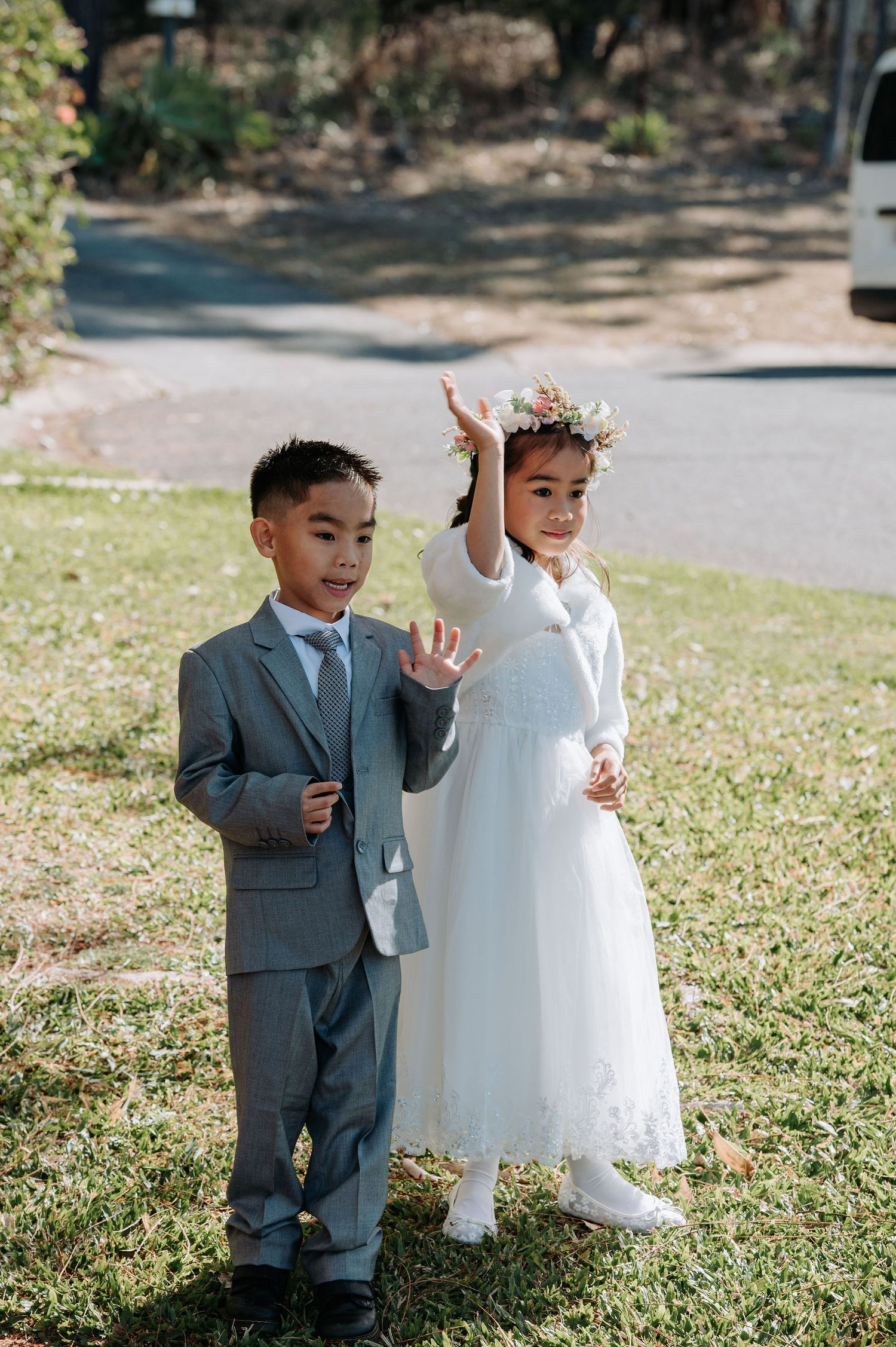 Two children in formal attire pose cheerfully on a sunny lawn.