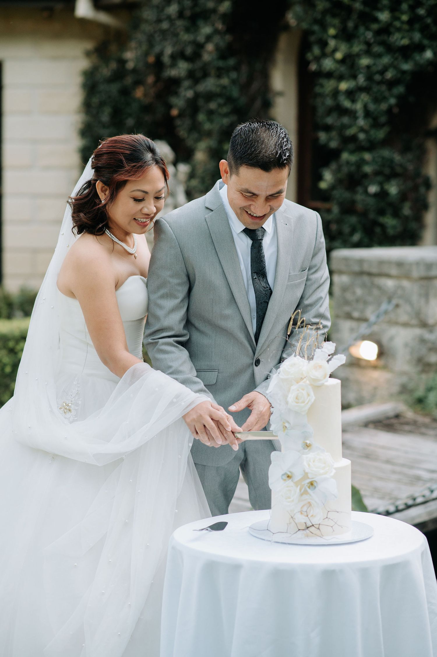 A couple cutting their elegant white wedding cake decorated with delicate floral details at their outdoor reception.