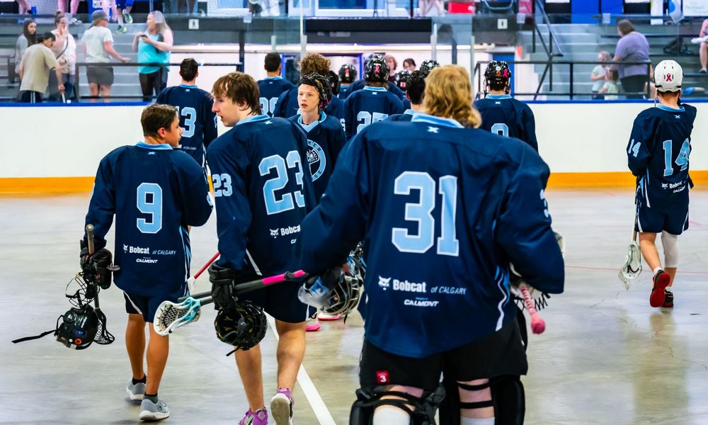 Lacrosse players in navy jerseys walking across indoor sports venue.