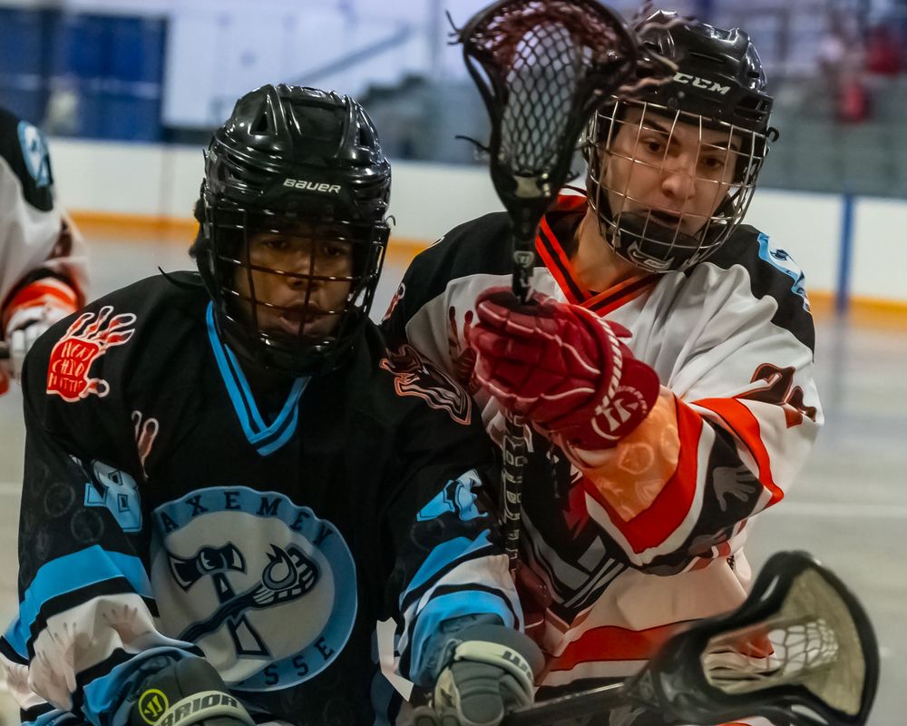 Two box lacrosse players in protective gear battle for possession during an indoor game.