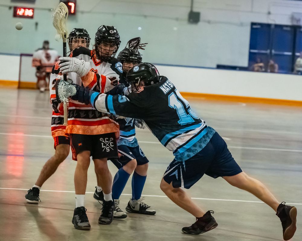 Box lacrosse players collide during an intense indoor match between blue and orange teams.
