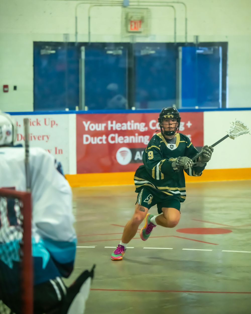 Player in green uniform carrying lacrosse stick while running across indoor arena floor.