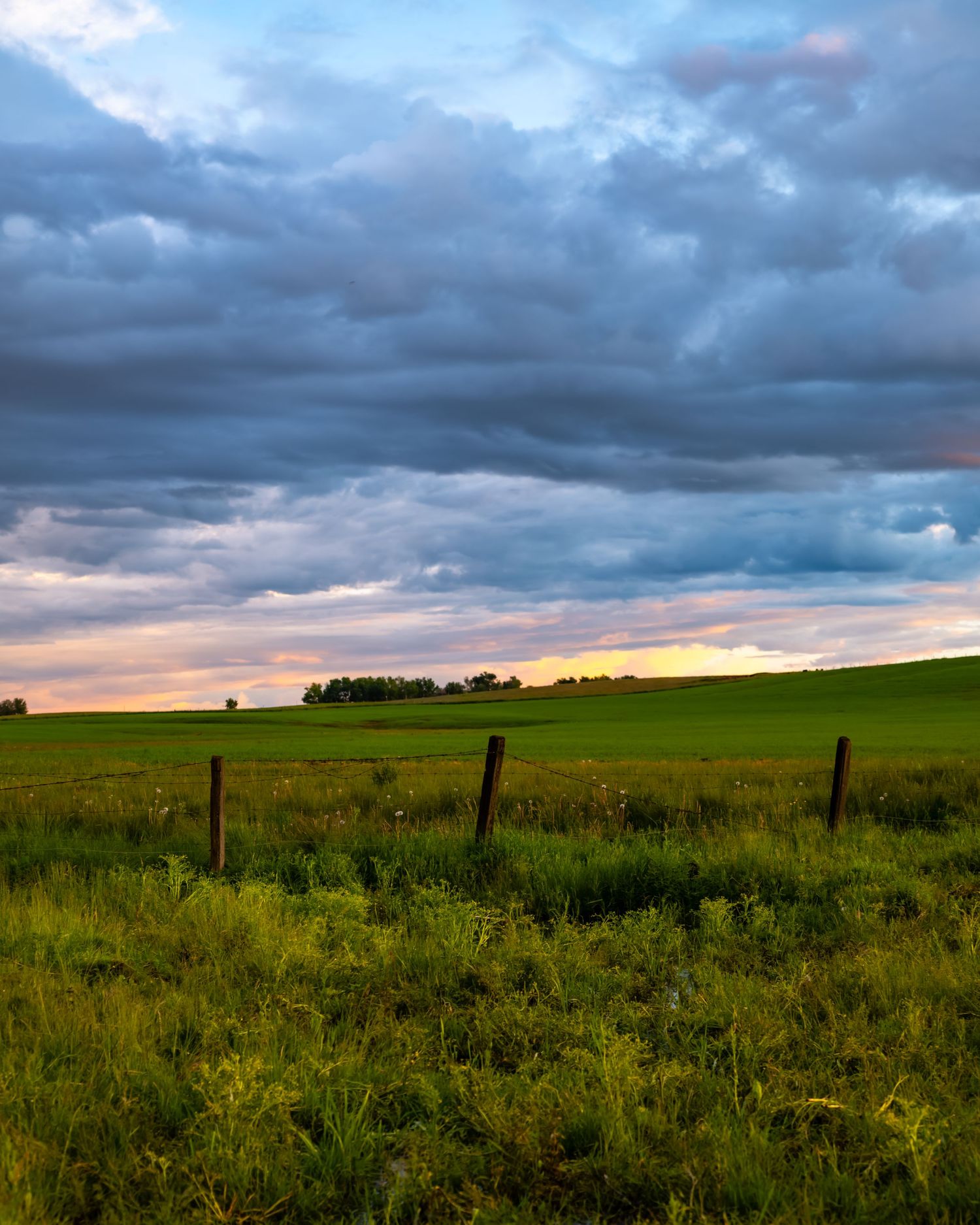 Dramatic storm clouds hover over a green rural landscape with wooden fence posts at sunset.