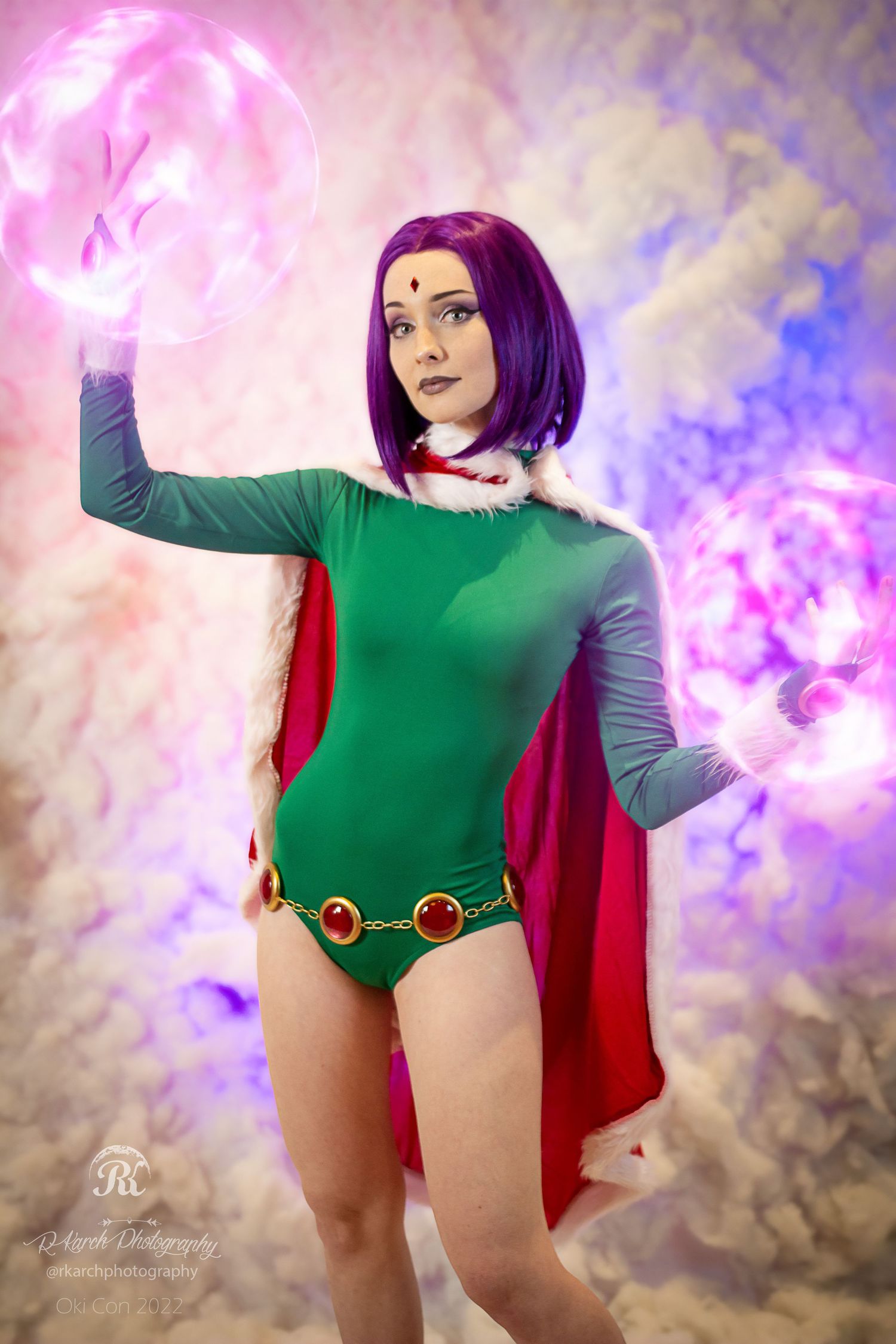 Cosplayer in green bodysuit with red cape poses with glowing pink magical effect.