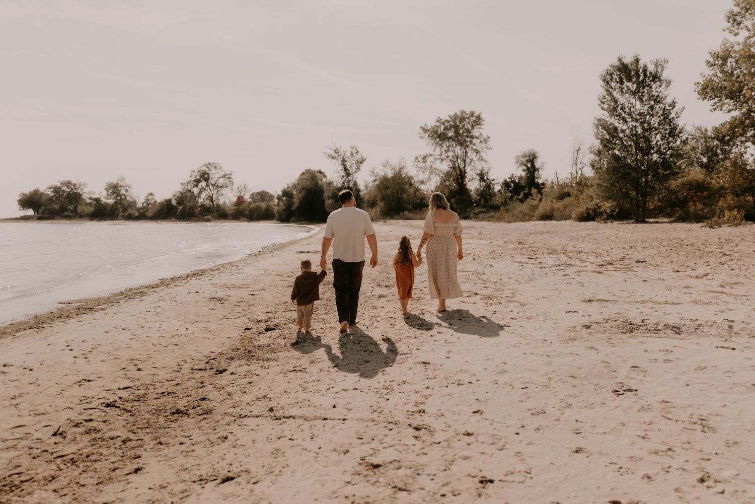 Family of four walking hand-in-hand along a sandy beach shore surrounded by trees on a sunny day.