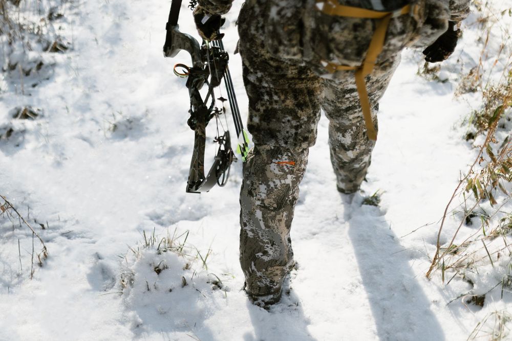 Half-body view of legs of person in camo clothing walking through the snow holding a bow and arrow