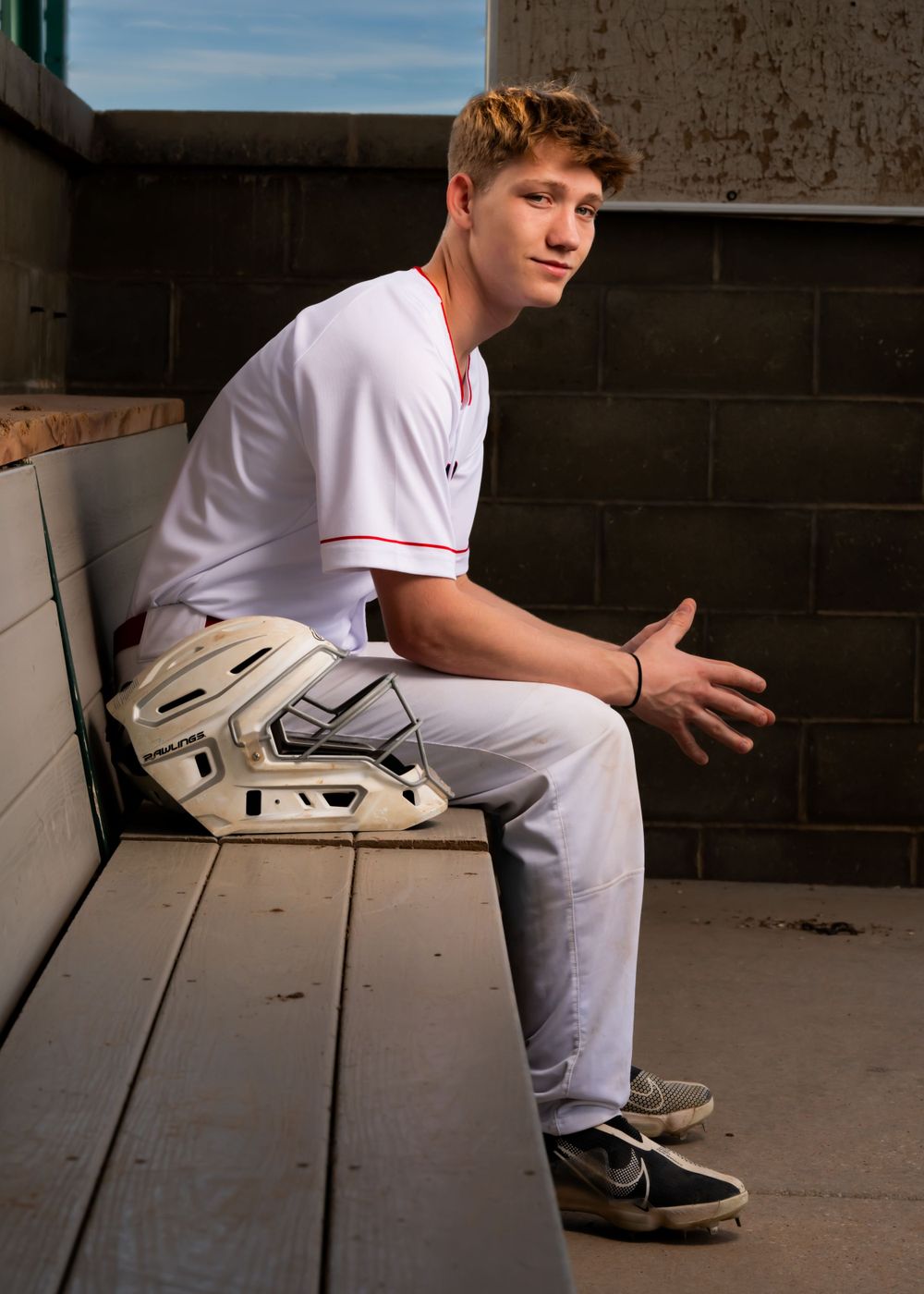Baseball player in white uniform sits in dugout with protective gear.