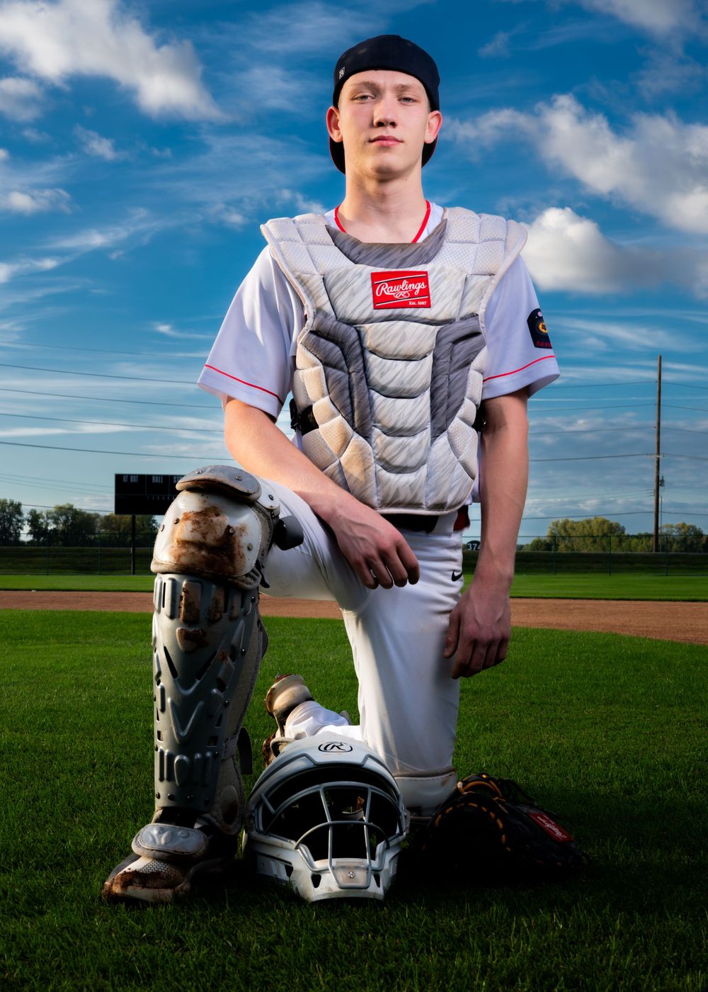 Baseball catcher in uniform kneels on field with protective gear under blue sky.