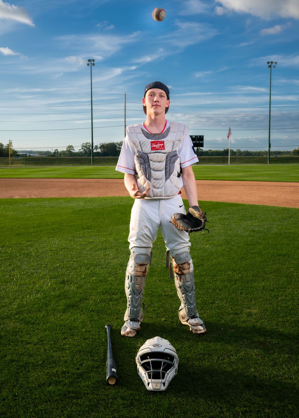 A baseball player in white uniform tosses a ball on a field with equipment at their feet and stadium lights overhead.