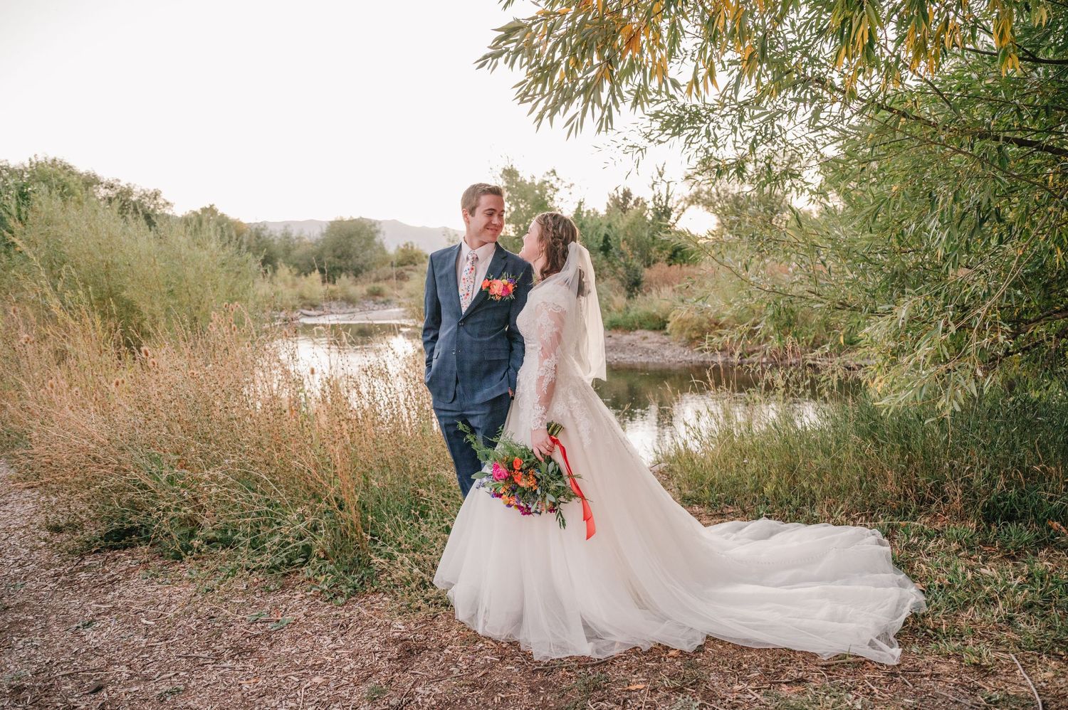 Newlyweds sharing romantic moment by waterside with bride holding vibrant bouquet.