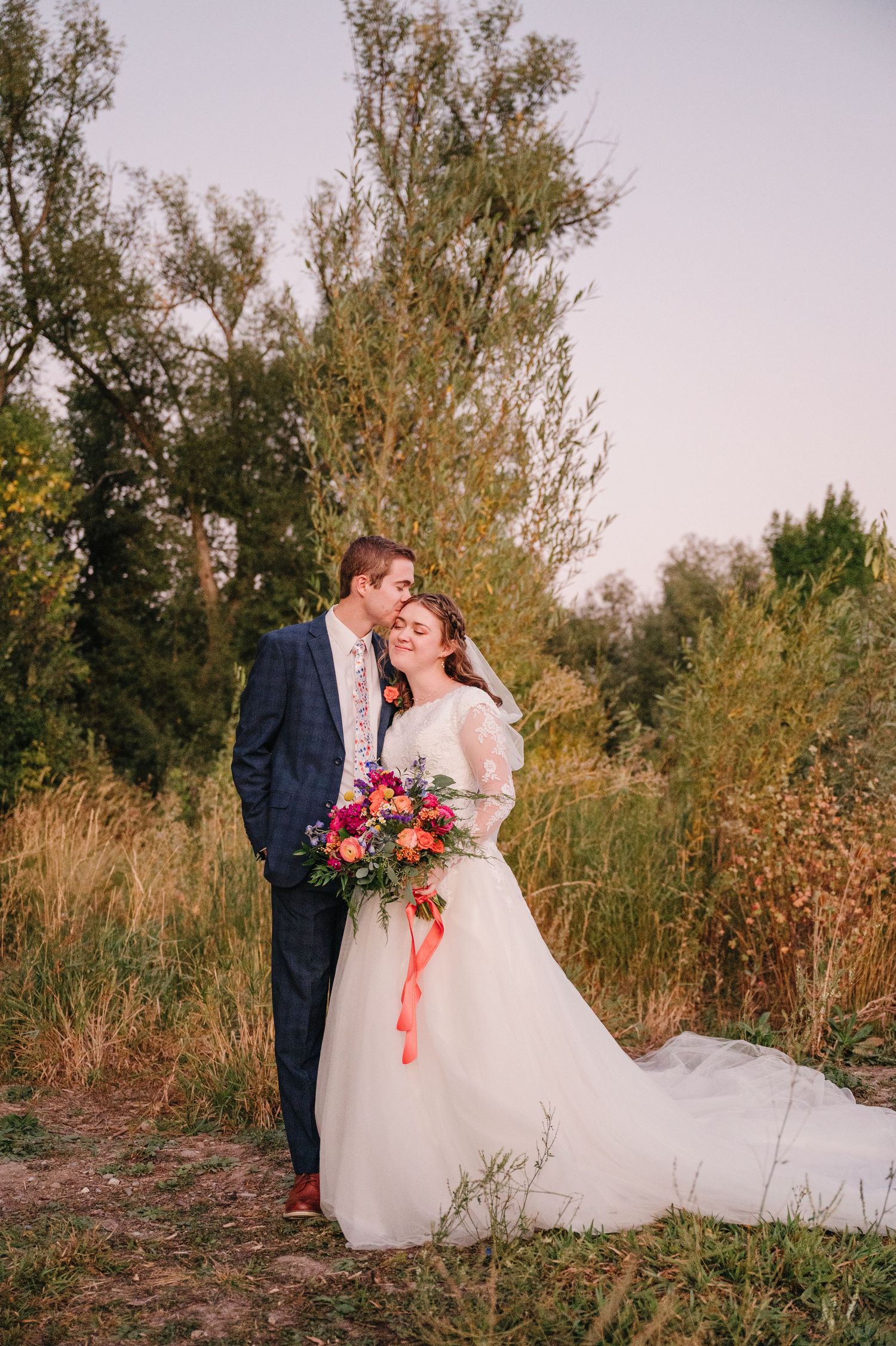 Romantic wedding portrait of couple with flowing white dress and colorful bouquet at sunset.