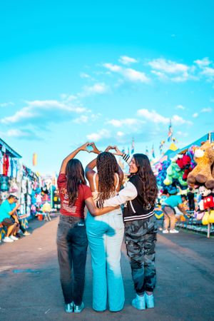 Three friends making heart shapes with their hands against a bright blue carnival sky.