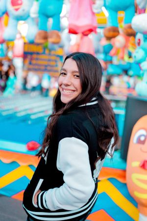 Person in varsity jacket smiling in front of carnival game booth with colorful plush prizes.