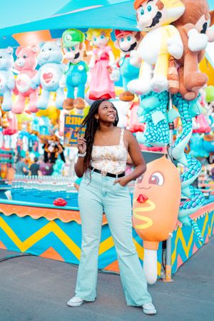 Person posing in front of vibrant carnival game stall with plush toys and decorations.