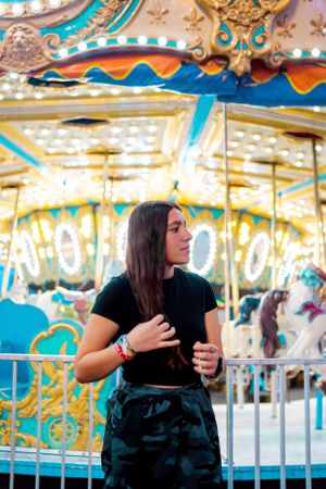 Person in casual wear posing by ornately decorated carousel at carnival.