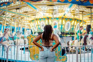 A vibrant carousel at a carnival illuminated with colorful lights as someone stands in front of the metal railing.