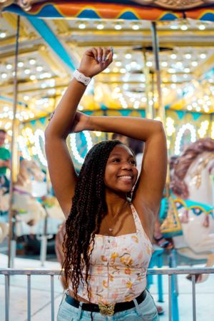 Young woman in floral top and jeans dances joyfully in front of a brightly lit carousel at a carnival.