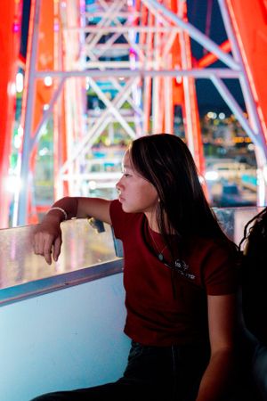 Person in burgundy shirt gazes at carnival lights while leaning on railing at night.