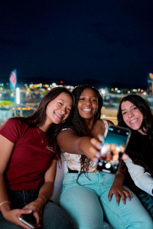 Three friends taking a selfie together at night with carnival lights twinkling in background.