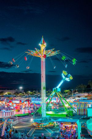 Vibrant illuminated carnival swing ride against dark blue sky at night with colorful lights and attractions below.