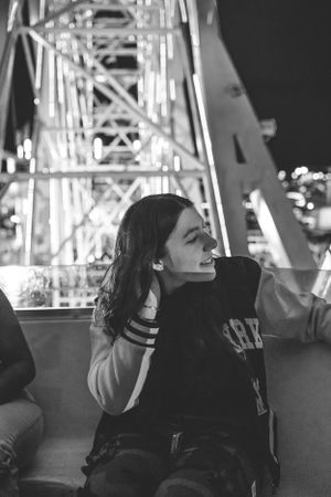 Black and white photo of person at a carnival with ferris wheel lights in background.