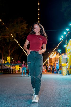 Person in red shirt and jeans poses under string lights at a nighttime carnival.