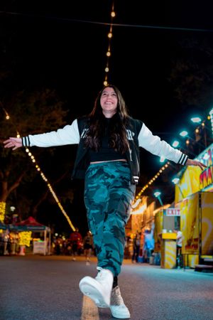 Person in camo pants and varsity jacket poses joyfully at nighttime carnival.