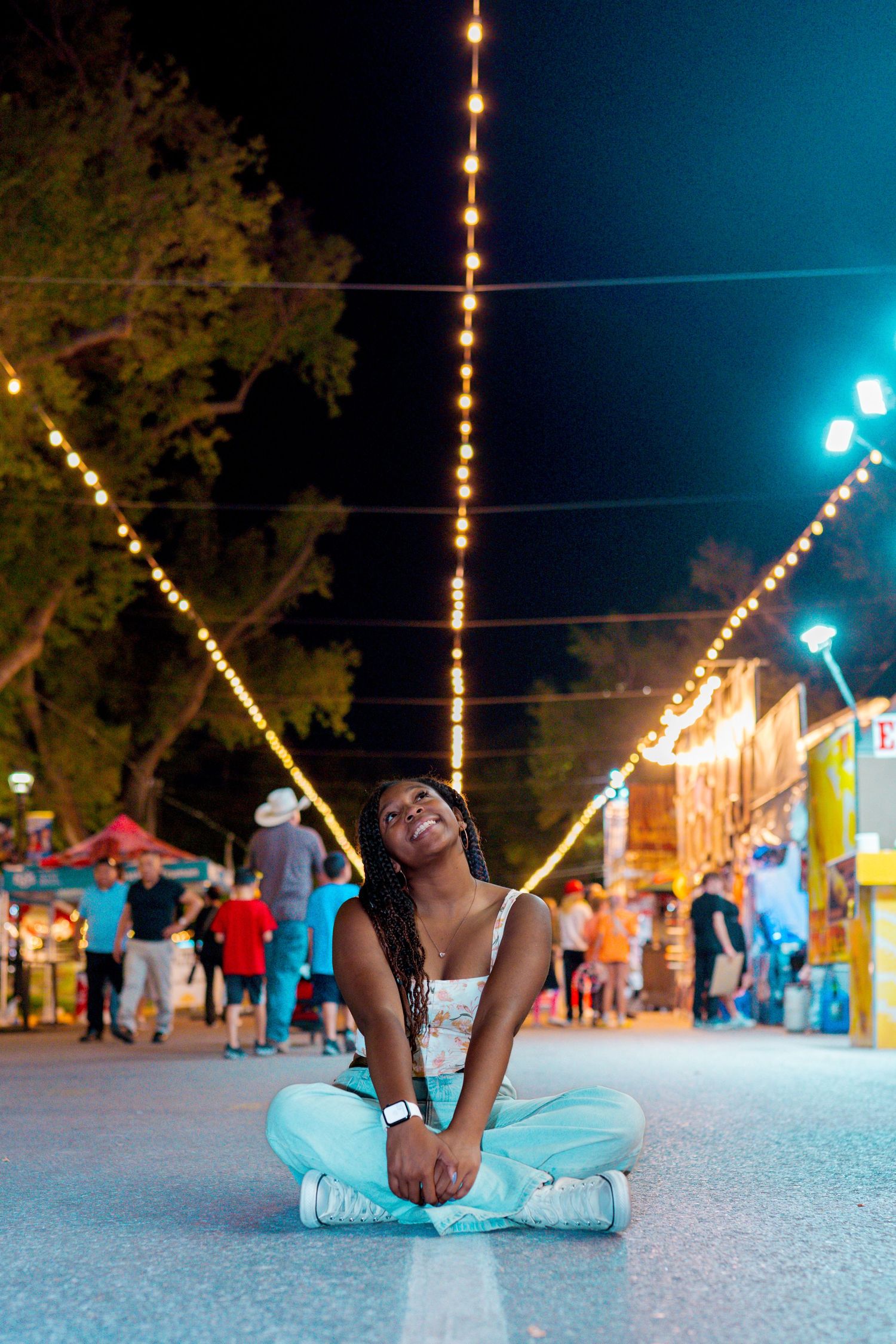 Person sitting cross-legged on carnival ground under string lights at night.